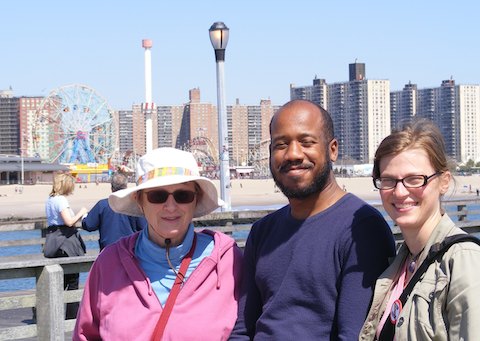 A close up photo of Gail, Eric and CJ with the 
             ferris wheel and other rides visible beyond the
             water and sand. Behind the amusement park are high-rise
             buildings.