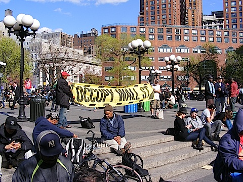 Two men hold a
        yellow banner proclaiming Occupy Wall Street. Two men in
        the foreground play chess. The sky is a beautiful blue.