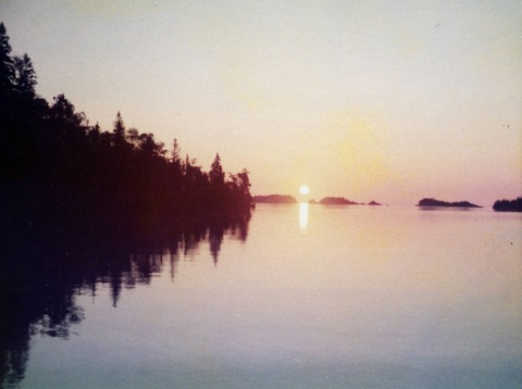 The sun is seen risen over the water with a tree-covered
              shoreline silhouetted to the left and some small islands 
              silhouetted to the right.