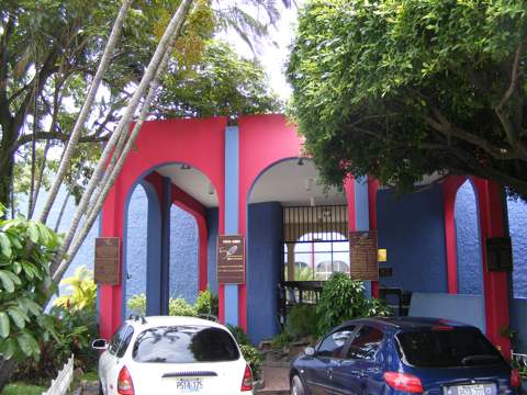 An entrance into the red and blue painted building, 
                    with cars parked in front and trees