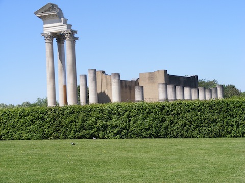 Majestic columns, mostly incomplete, supporting
             part of a portico, rise behind a green hedge