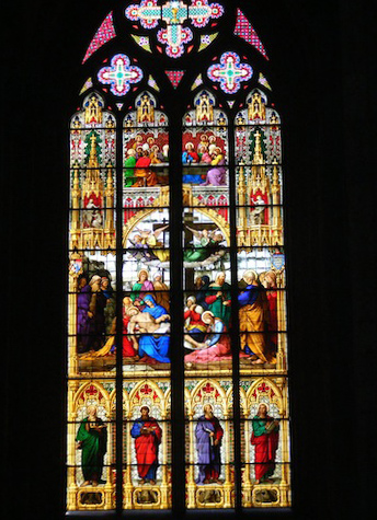 This large colored window incorporates many images. At the
           bottom in an area four panels wide and three high are representations
           of the writers of the four gospels standing in archways. Each
           has a representative animal at his feet. Above them in an area
           four panels wide and four high is a representation of the body
           of Jesus being removed from the cross. Above that is a two by two
           picture of people around a table, possibly a representation of 
           the last supper. Above that is an area with two smaller and one
           large cross, mostly in the colors of lavender, rose and white.