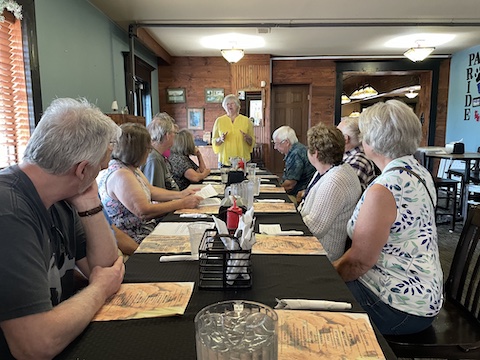 Eunice is standing at the far end of the table with the other
             attendees sitting on bothh sides of the table and looking at her