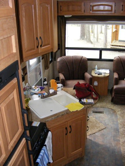 The two-basin sink and its cabinet sticks out
          into the living area at about a 45 degree angle. A small amount of
          the range and the refrigerator can be seen at the left side. 
          The two easy chairs are visible near the window at the back of the
          room.