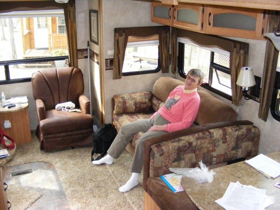 Gail is sitting
          on our little couch on the side of the living
          area. One of the easy chairs is visible at the
          back of the room.