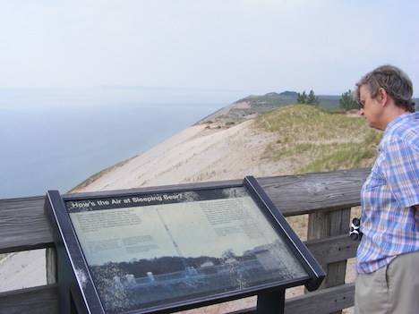 Gail reads a plaque on a viewing
                platform. A steep sand and
                rock slope stretching from the top of the
                dunes down to the lake shore can be seen
                behind the plaque 