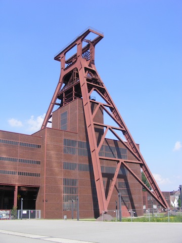 A barn red A-frame tower straddles a rusty red building in front of
              a deep blue sky