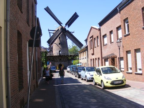 At the end of a narrow street, a 17th century windmill stands.
            The street is lined with brick buildings and cars are parked along
            one side. 