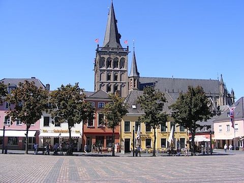 Across a wide, paved open area stand a line of colorful shops. 
                Rising high above the shops is the bell tower of the cathedral.