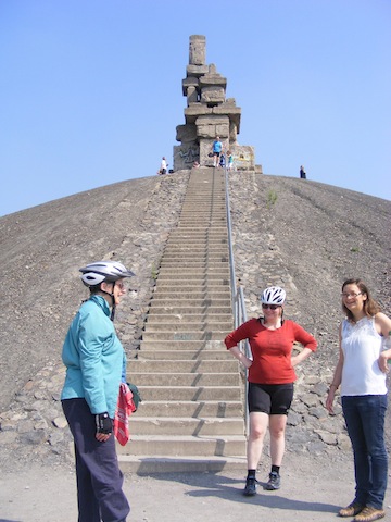 Gail in a blue shirt and bike helmet, Ingrid in a red shirt and
             bike helmet and Katrin in a white shirt are standing at the bottom
             of a long stairway that ascends a hill of rocks. At the top of the
             hill is a sculpture that consists of massive blocks stacked into
             a tower that is about 30 feet high. 