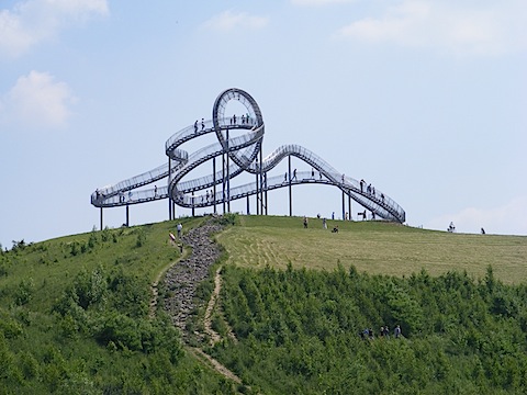 At the top of a grassy mound is a curlicue structure, maybe 130 feet
          across and 40 feet high. People are walking on the pathways formed by
          the structure.