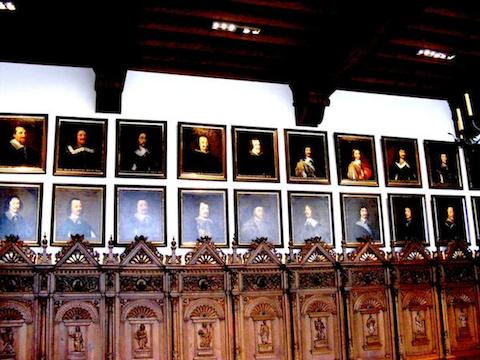 Twenty portraits of men in two rows are seen. Most of the men are
             wearing black garments with a collars. The young Louis XIV of France 
             has a gold and crimson garment. Below the portraits can be seen
             the ornately carved wooden backs of seats that line the wall.