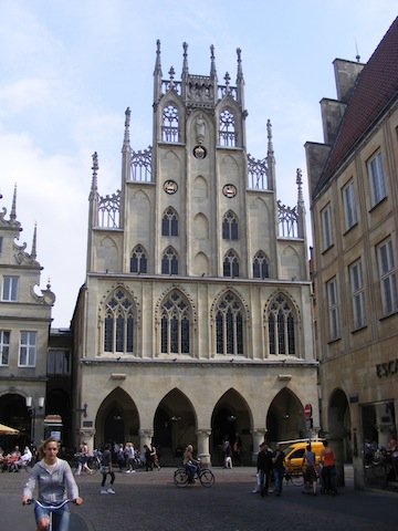 The facade of this building consists of four large open pointed arches
           at street level. Above them are four windows in pointed arches of about the 
           same size. Above them is a front that gets narrower in four square steps to
           the top. In this false front are six windows in pointed arches as well
           as decorative arches with solid interiors. At the top are two statues.
           The whole is made of tan stones.