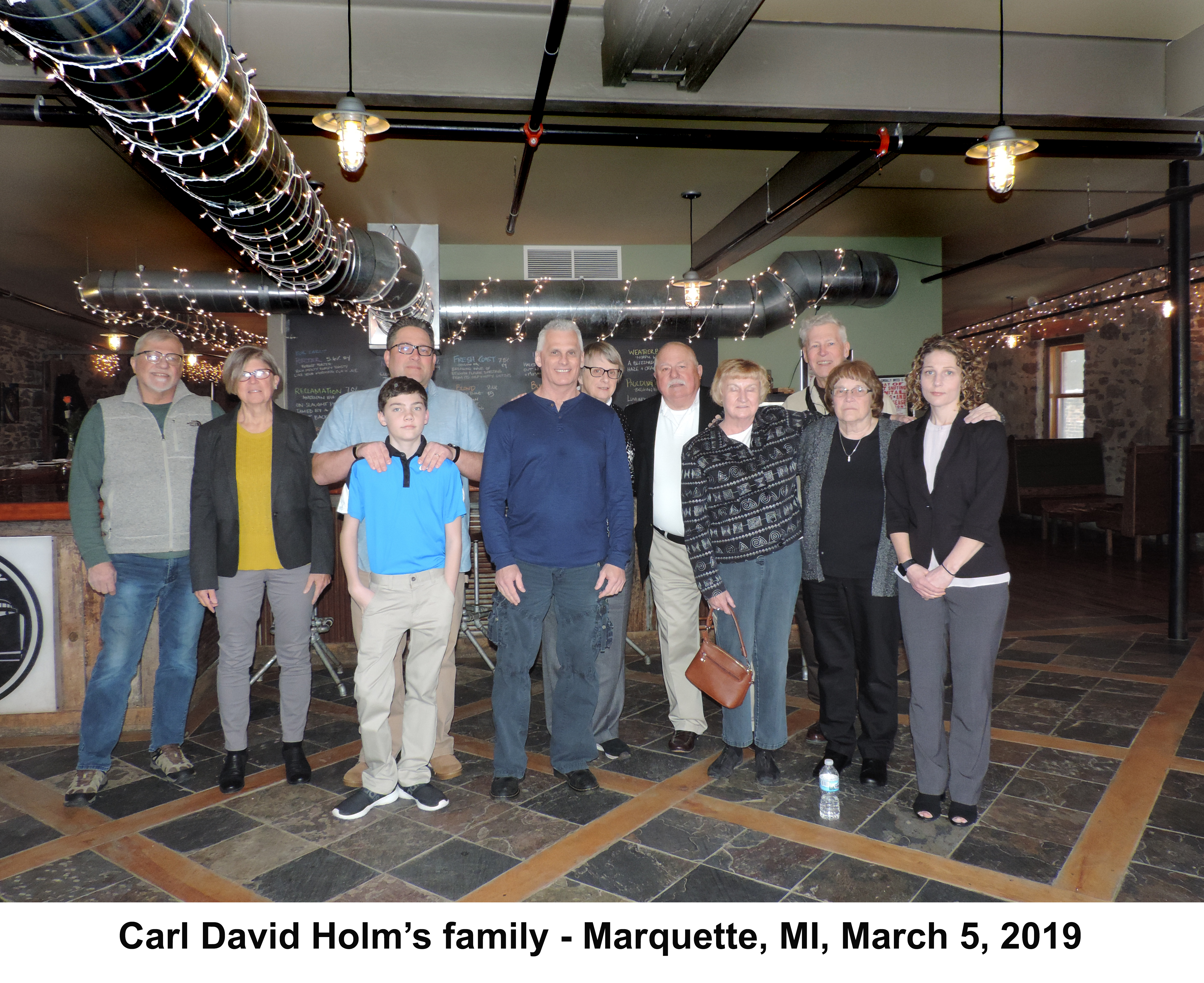 His siblings, cousins, and children with his wife Ruth Ann and 
           Albert's wife Gail are standing in a large room with heating ducts behind them.'