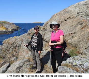 Britt-Marie and Gail are standing on the rocky coast with blue sea, blue sky,           and two rocky islands behind them.