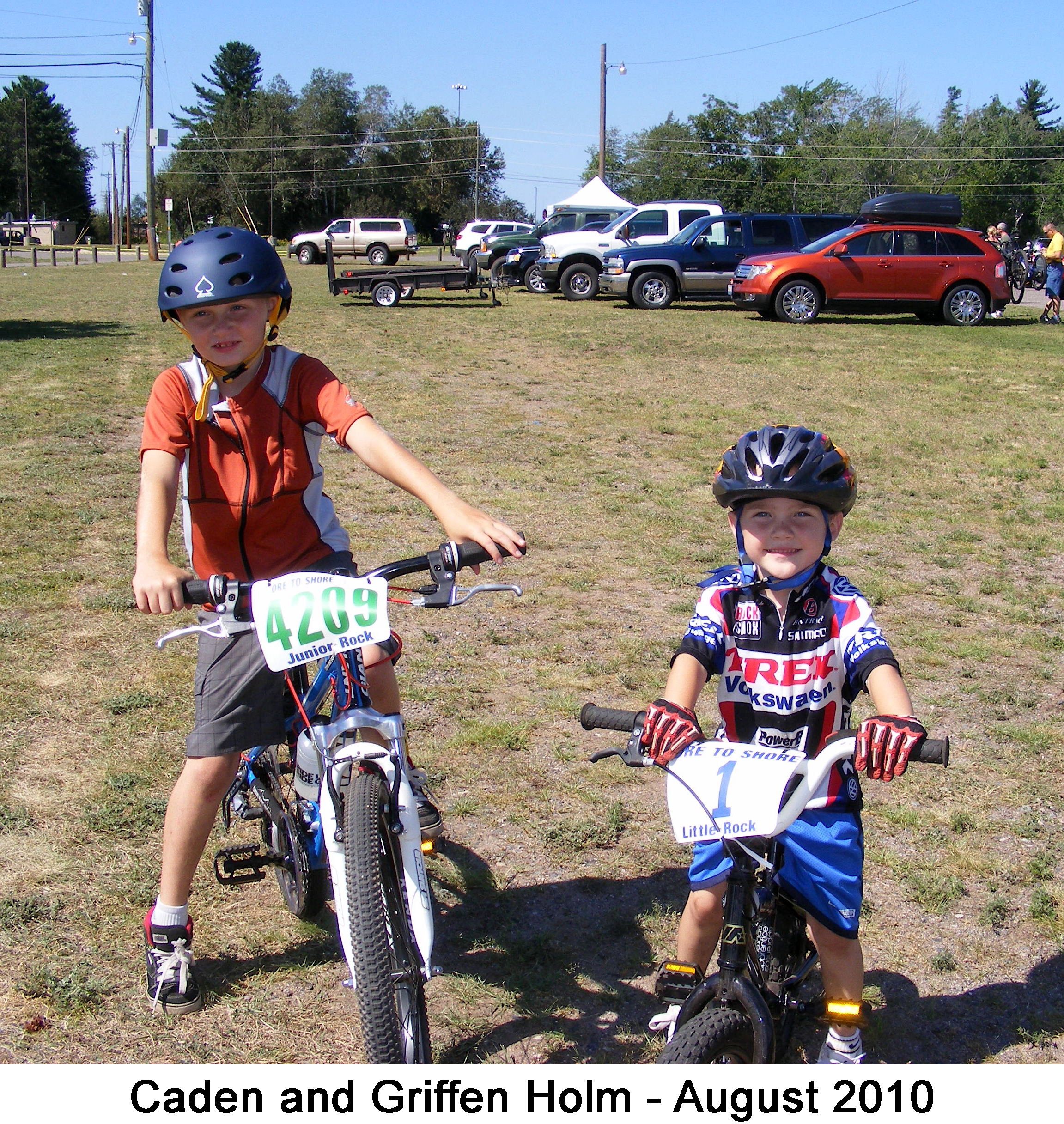 The boys are sitting on their bikes wearing helmuts and looking at the camera.
          Griffen has a big smile. There are cars in the background. 