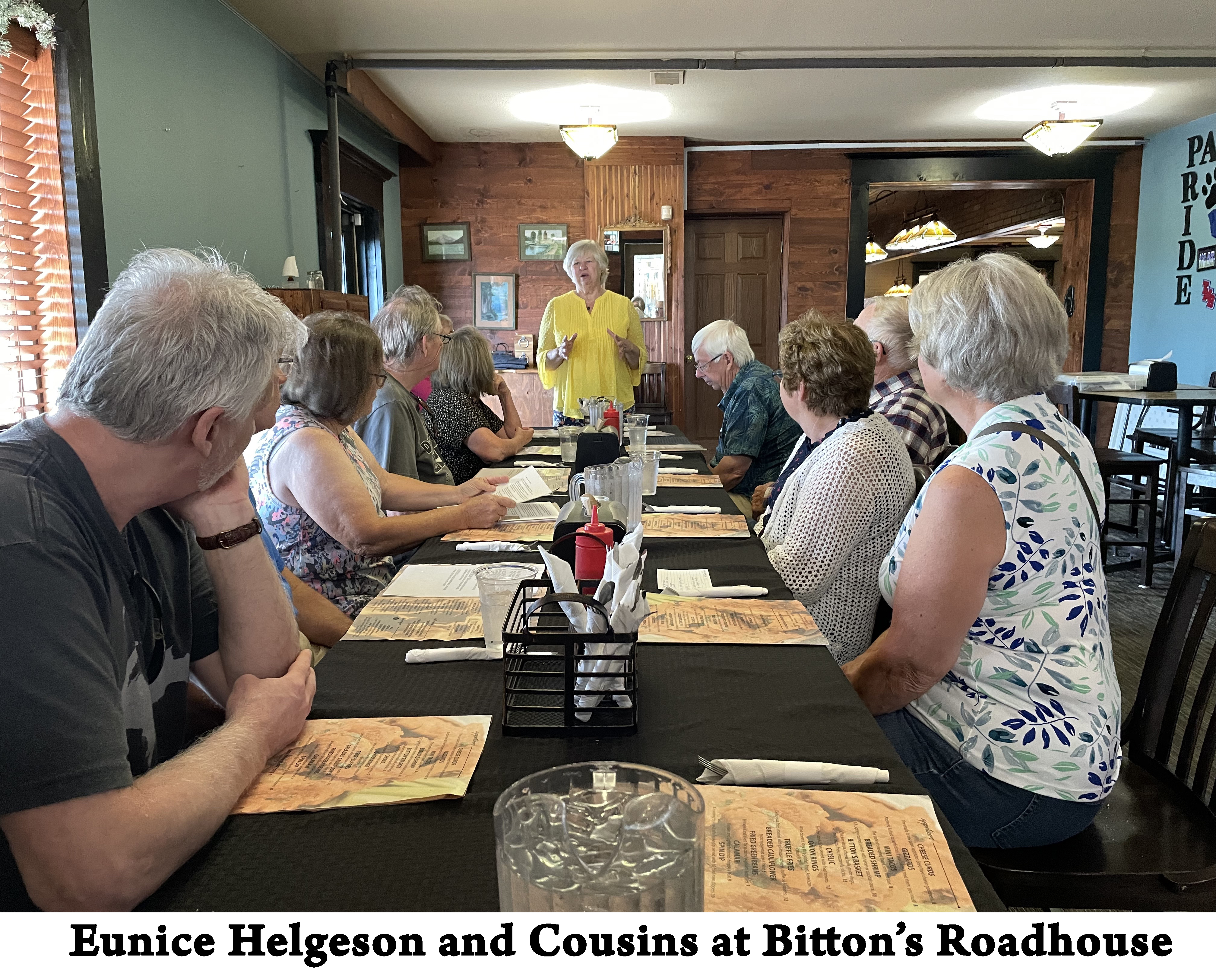 Eunice, wearing a yellow blouse, is standing at the other end of the long table. 