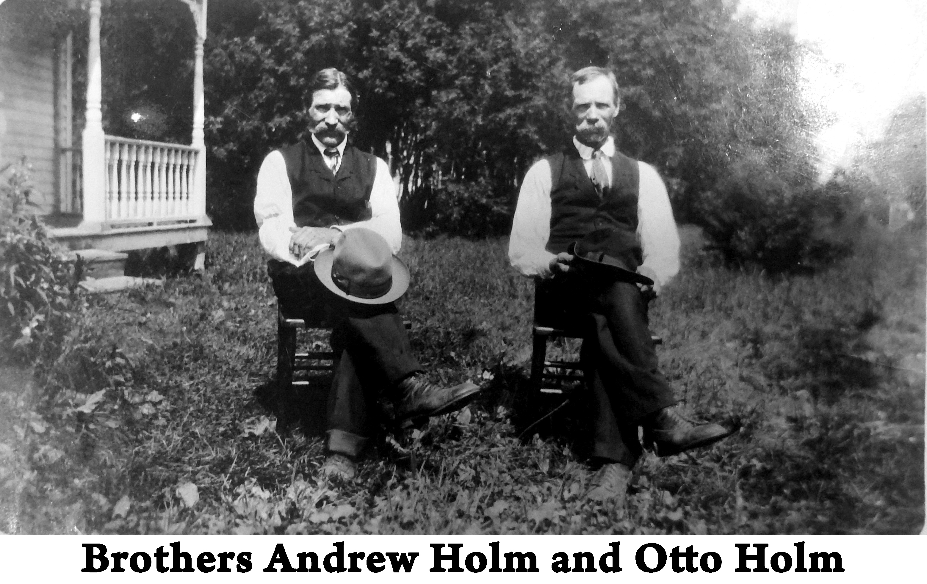 Two men are seated on kitchen chairs on the lawn in front of a house.
             Each is wearing a vest and a tie and holding a hat. 
             Both have mustaches. 