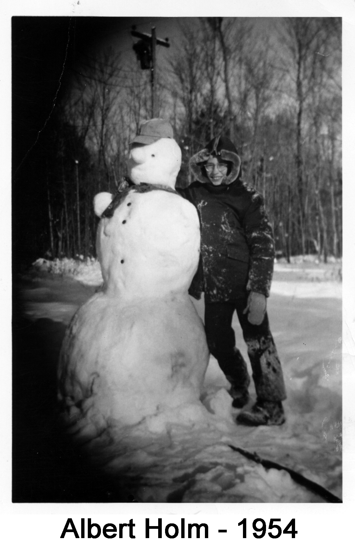 Albert in a hooded snowsuit is leaning against and hugging a snowman as tall as he is
