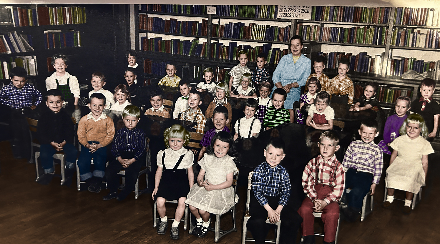A black&white photo of 35 students and their teacher in front of a wall of books.
       Most are sitting and smiling at the photographer. 