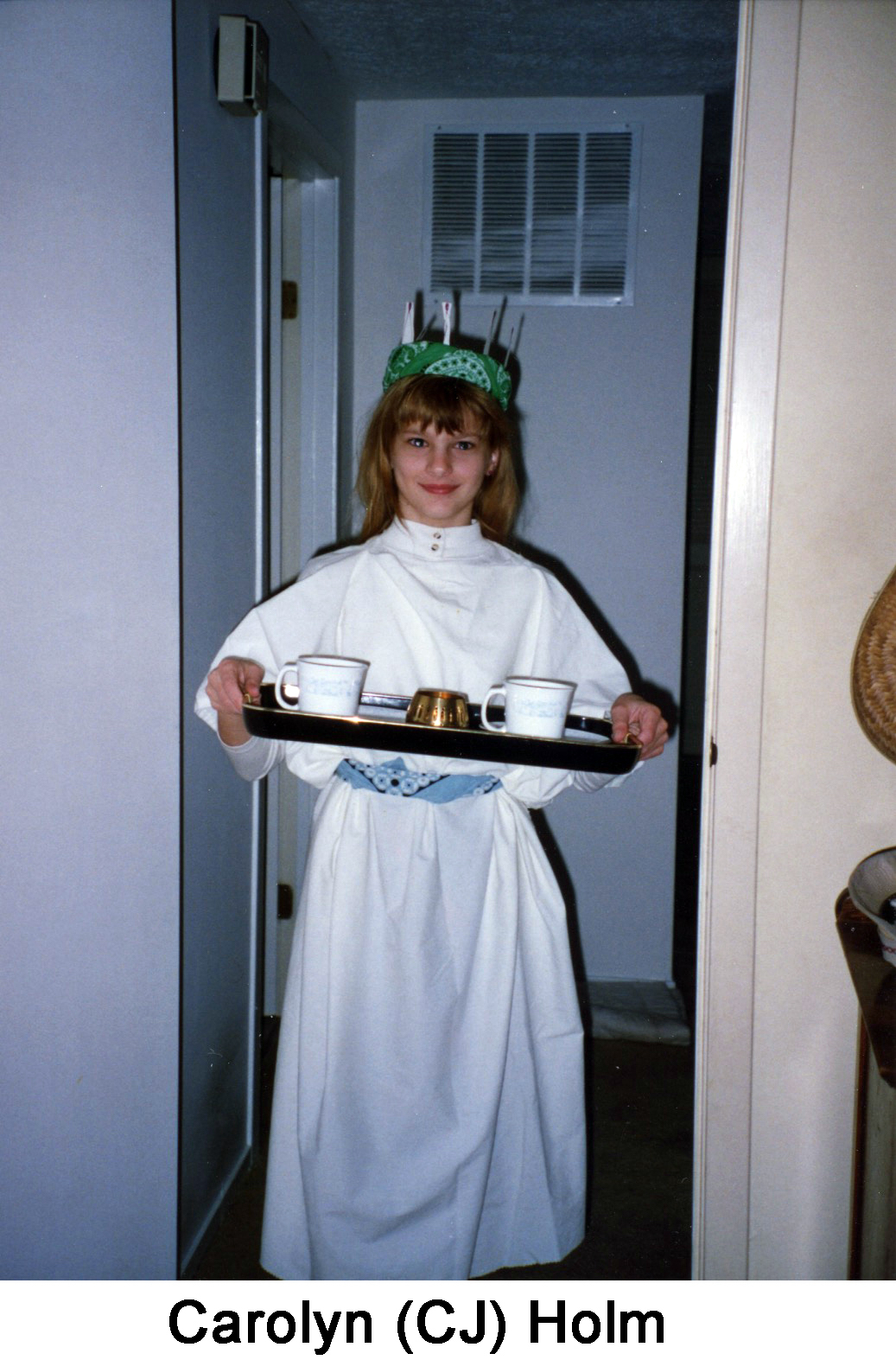 CJ is carrying a tray with two cups and wearing a crown with immitation candles.