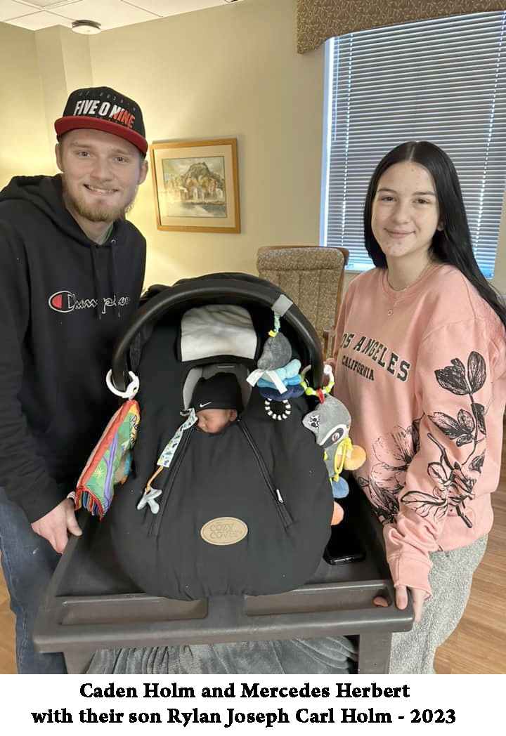 Caden Holm and Mercedes Herbert are looking at the camera with Rylan in a
             baby carrier between them 