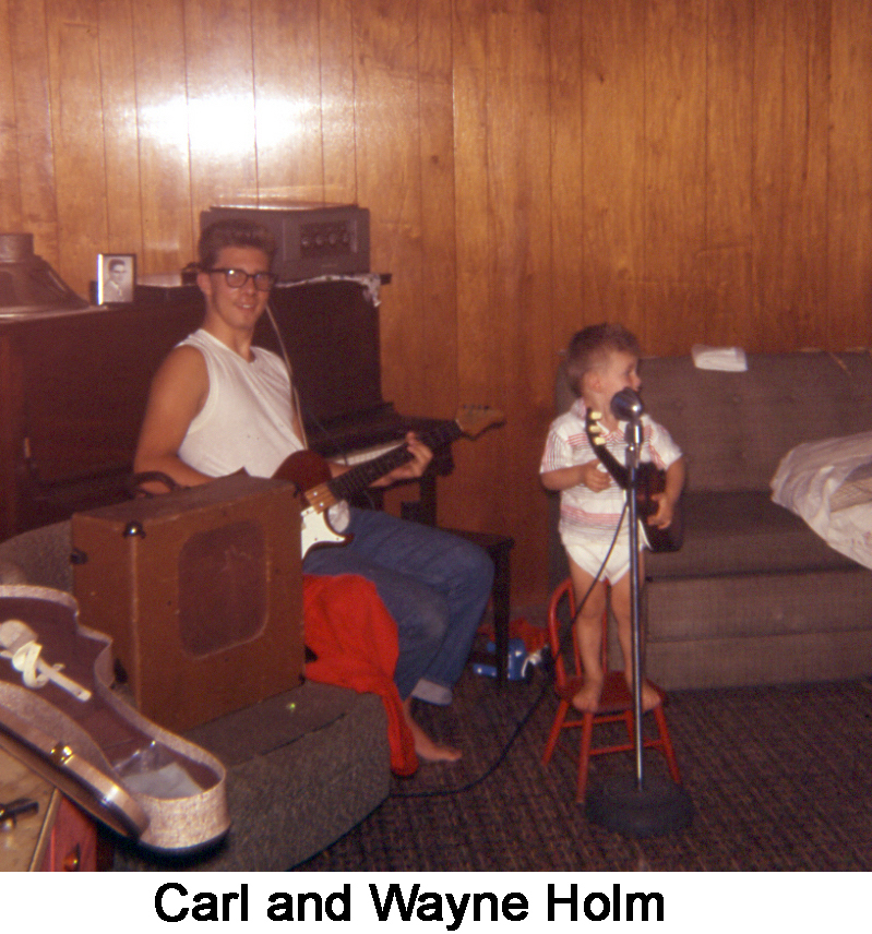 Carl is sitting, holding his bass guitar, and smiling at the camera.
        Wayne is standing on a child’s chair behind a microphone and holding 
        a ukulele. 