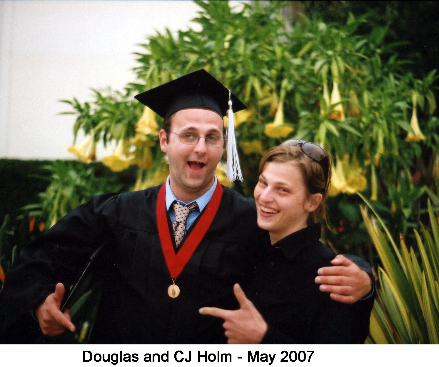 Doug is in his graduation robes with one arm around his sister. 
     A flowering plant is behind them. 