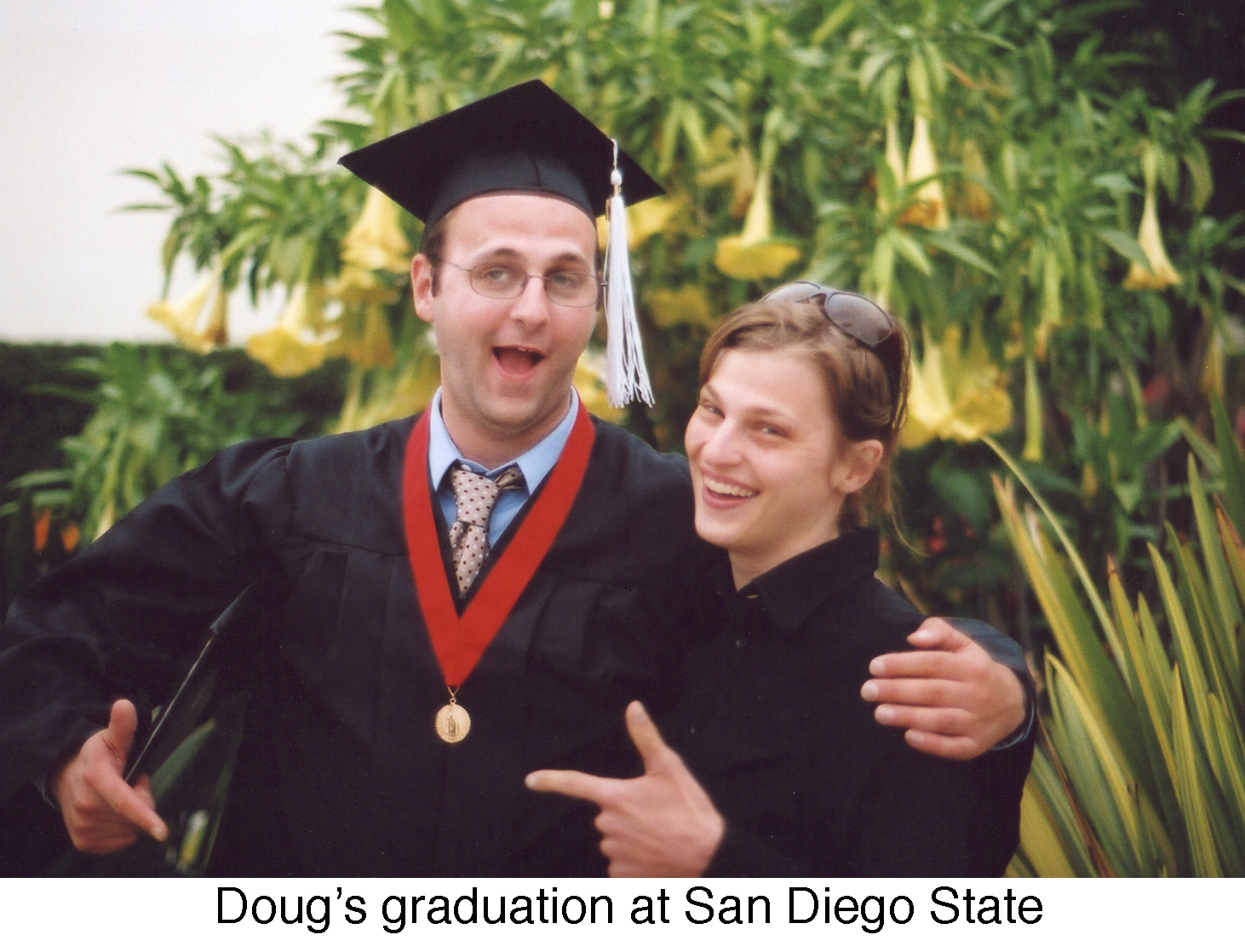 Doug is in his graduation robes with one arm around his sister. 
     A flowering plant is behind them. 