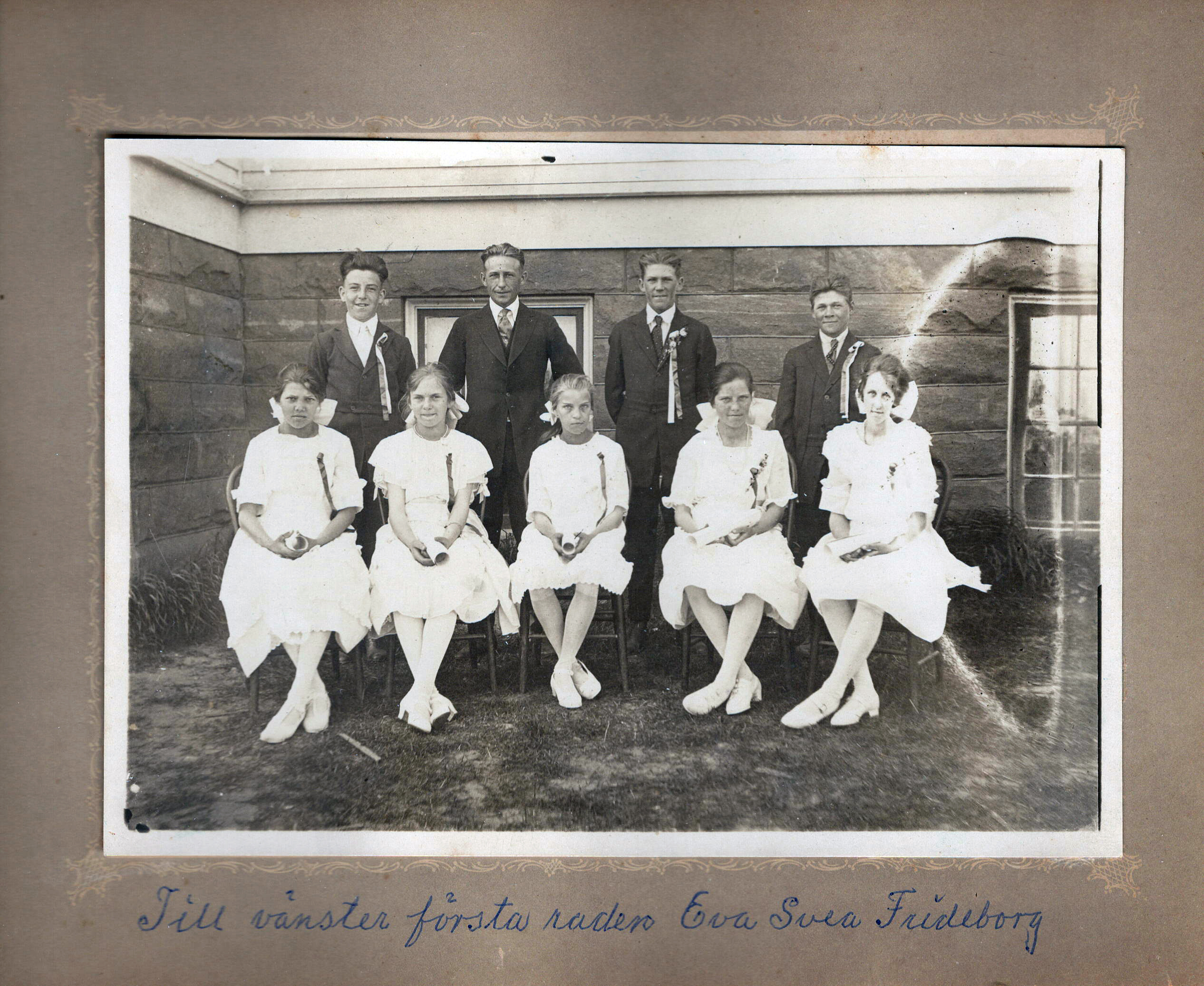 The girls are seated, and the boys and the teacher are standing behind them
        in front of the outside of the school. The photo is contained in a 
        cardboard frame.