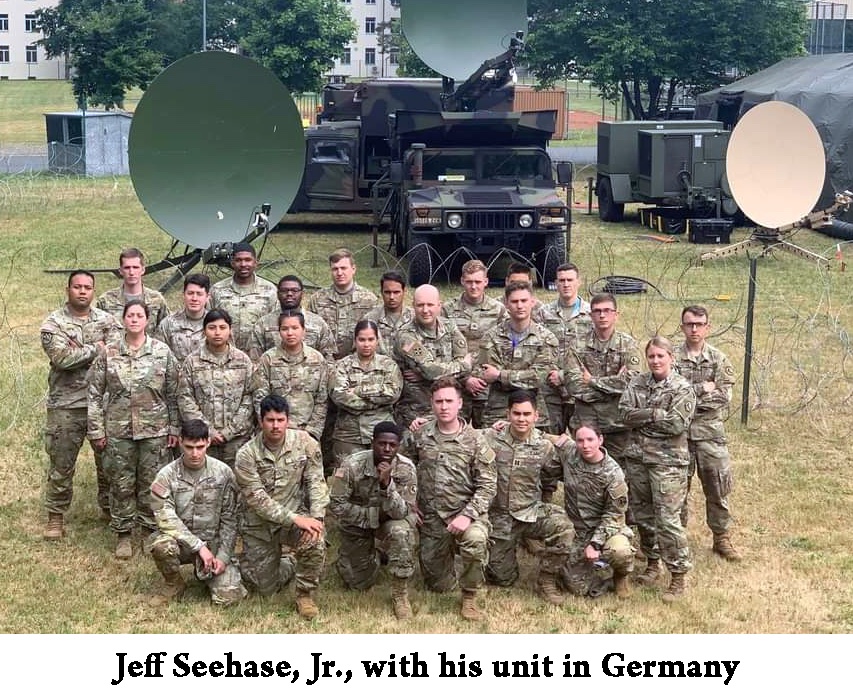 24 soldiers are standing in three rows in front of antennae and vehicles.