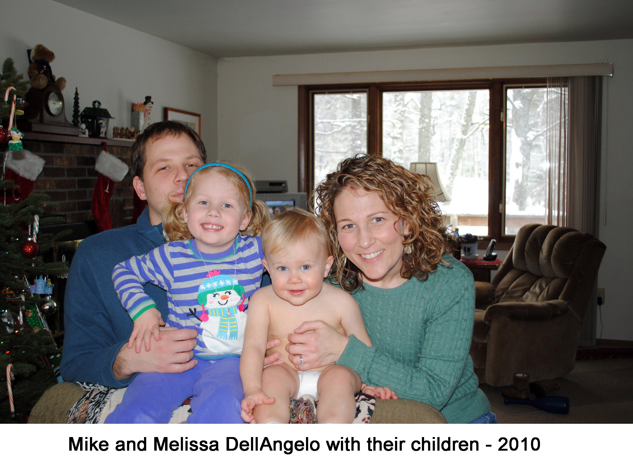 The family is sitting together with Mike holding Macyn and Melissa holding
     Evan. The snowy woods can be seen through the window behind them.