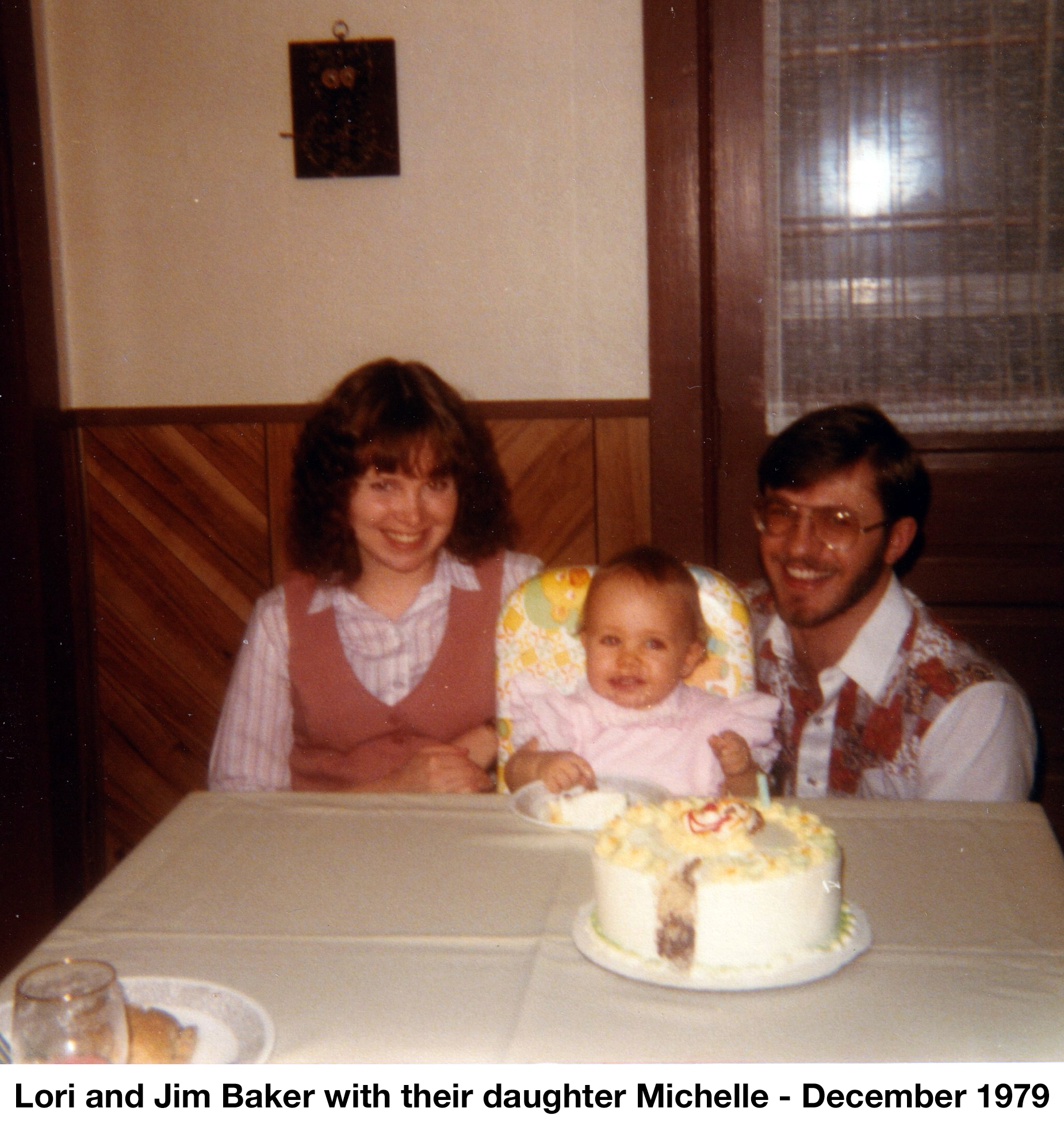 Michelle is in a highchair with her parents on either side and a 
         cake on the table in front of her