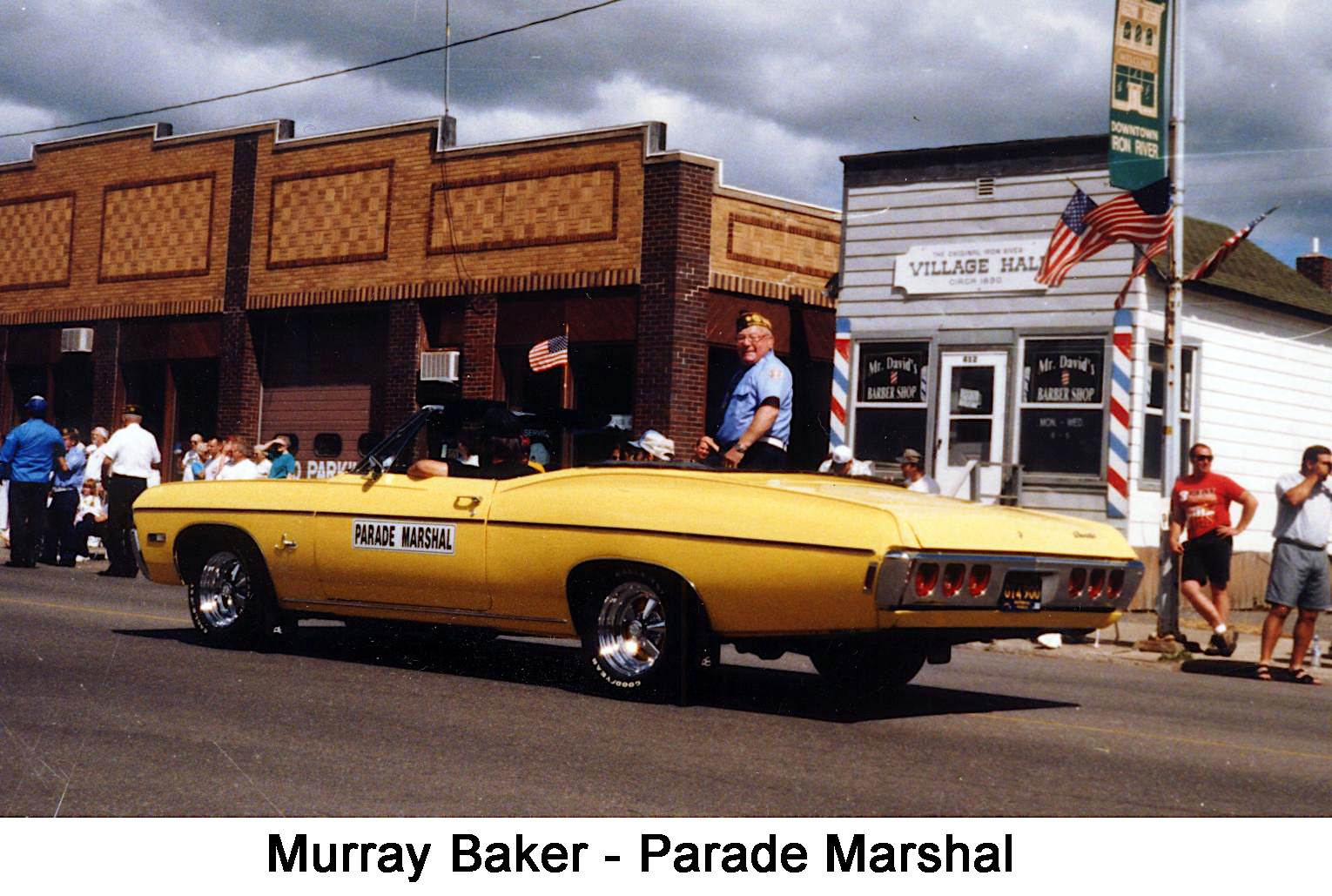 Murray is sitting on the back of a yellow convertible, which is 
     passing in front of the original Iron River village Hall, circa 1890, now 
     Mr. David's Barber Shop.