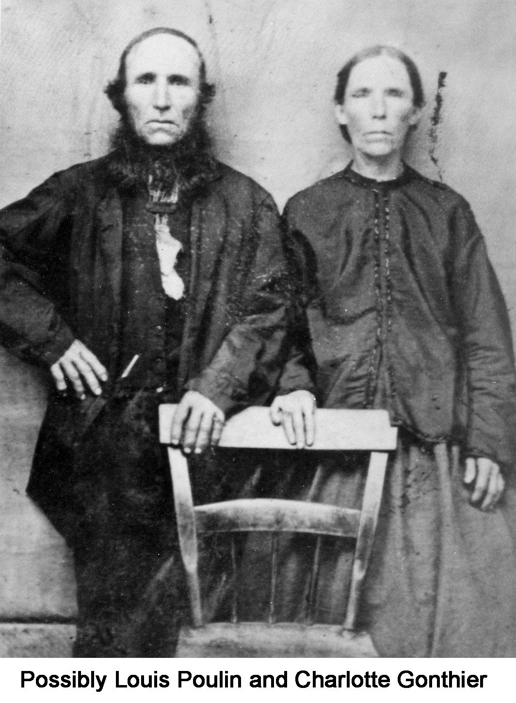 The couple are standing behind a kitchen chair and staring at the camera. 
         Both wear dark clothing. He has a beard, but no mustache. 
         Her hair is tied back.