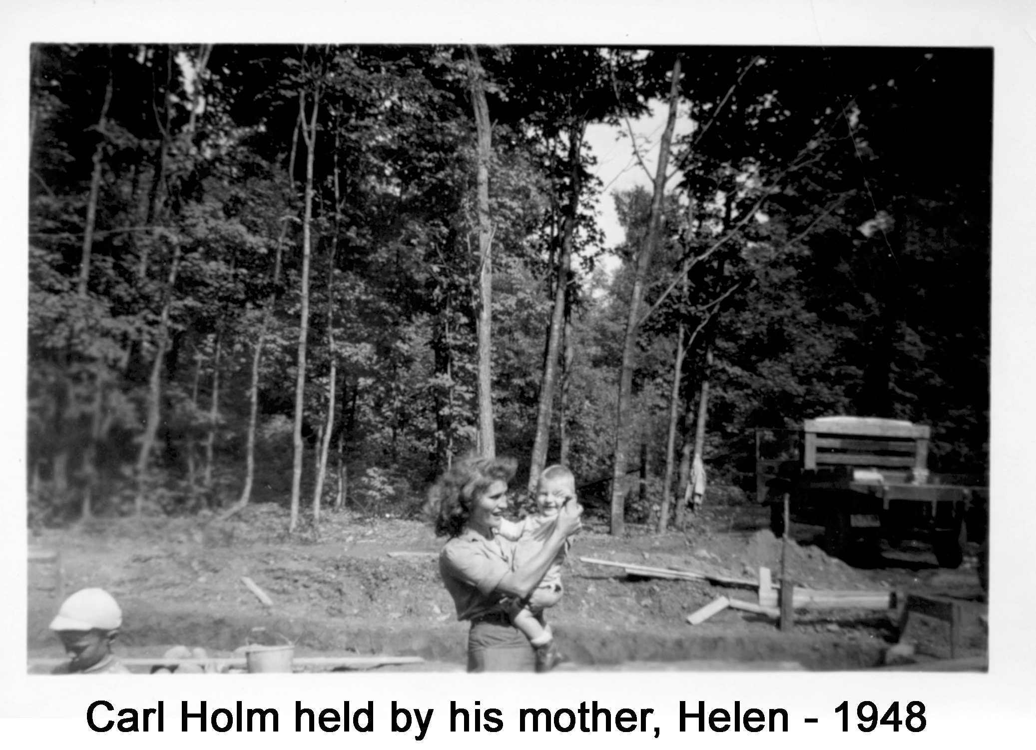 They are smiling. A truck is parked on the right. A line of trees is behind the excavation.