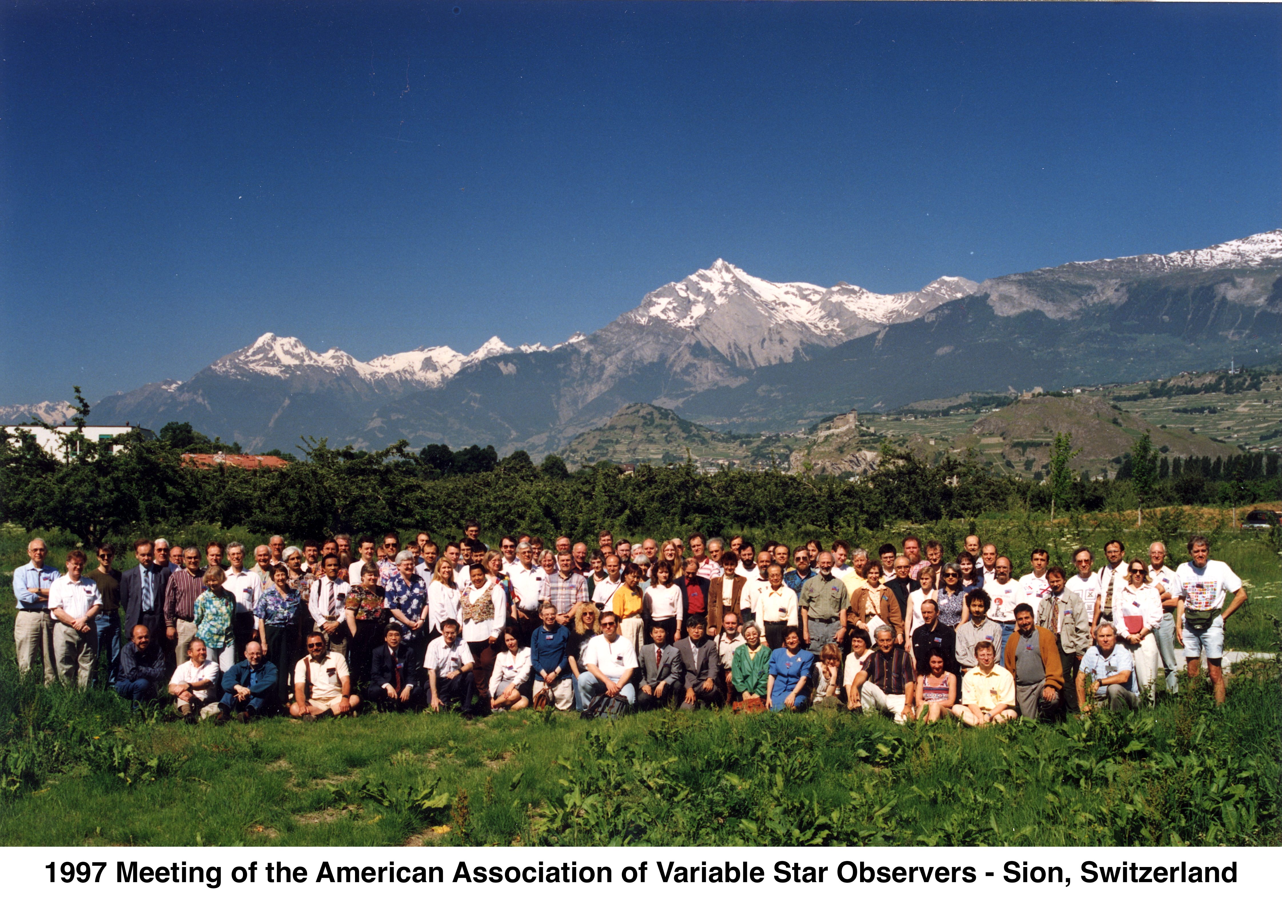 The participants are lined up in front of green shrubs with 
            snow-covered mountains and a clear blue sky in the background.