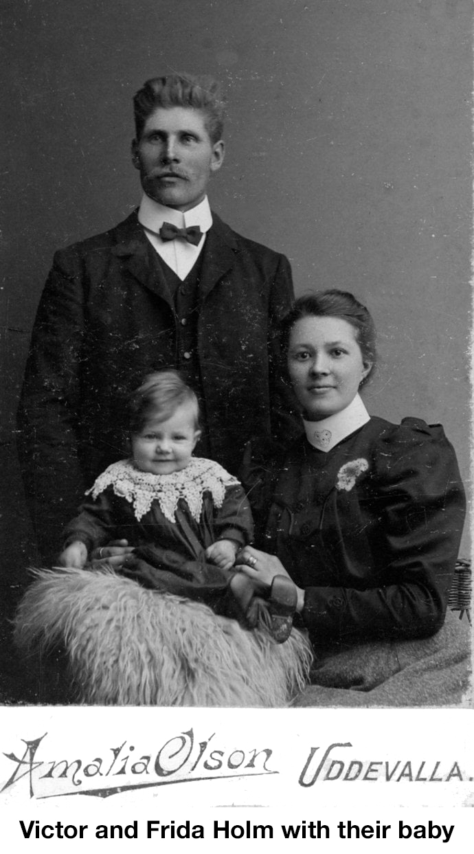 Victor is wearing a suit and tie, and standing. Frida is sitting to 
       his left. The baby is on a cushion in front of him.