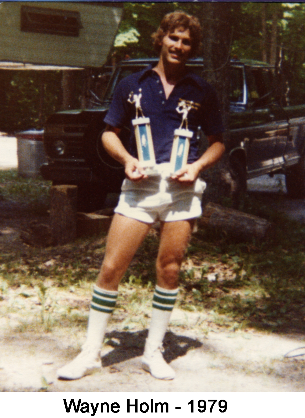 Wayne is standing in the yard in front of a pickup and holding two tennis trophies.