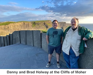 Danny and Brad are standing next to a wall. The cliffs are visible in the 
         distance. The sea is visible in the distance on the right.