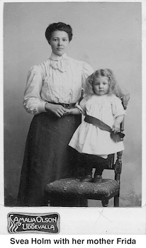 Svea has long, curly blond hair. She’s wearing a white dress and standing on a chair. Frida is standing beside her.