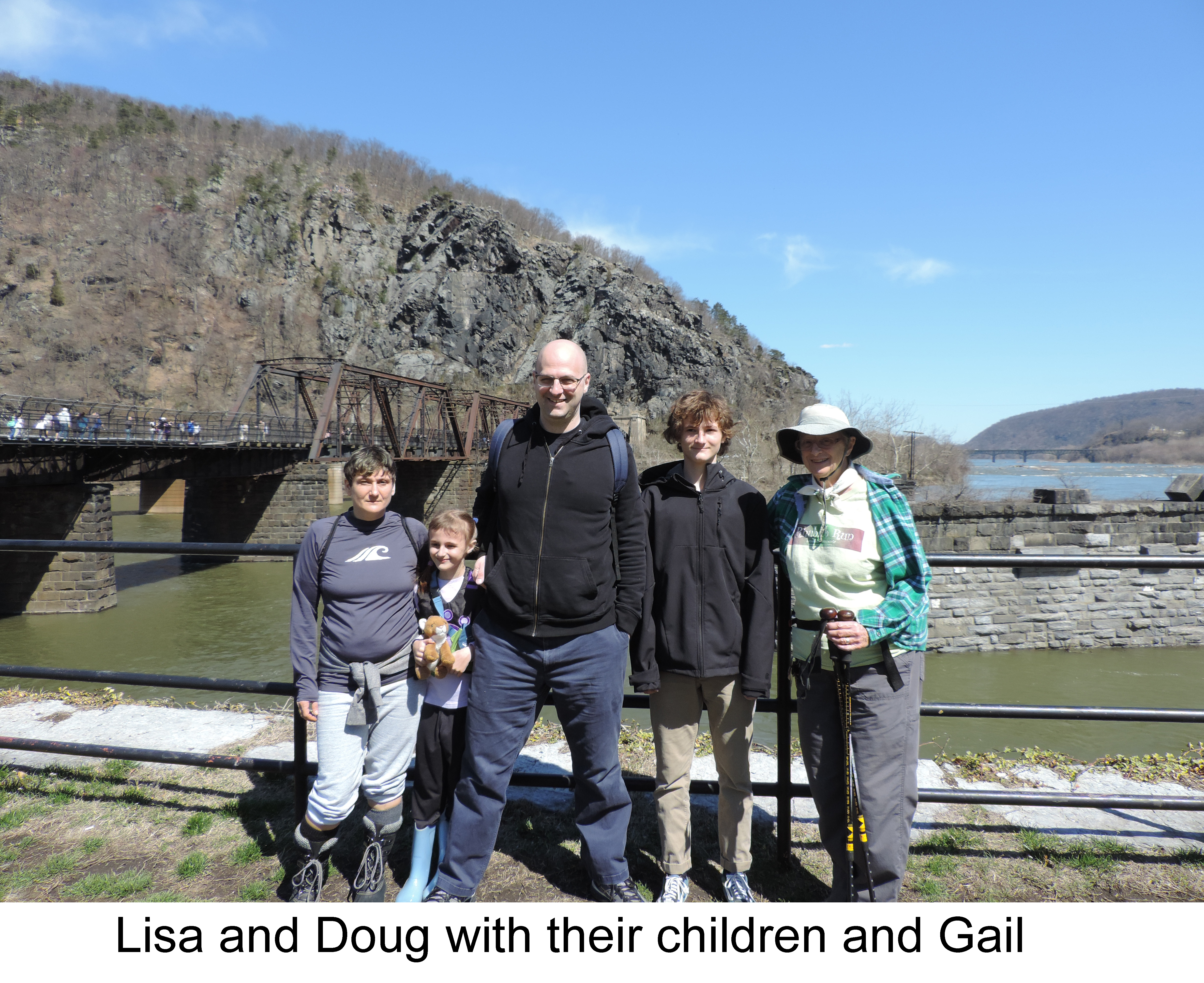 The family is standing in front of the river on a sunny day.      People are walking across the footbridge on the left. 