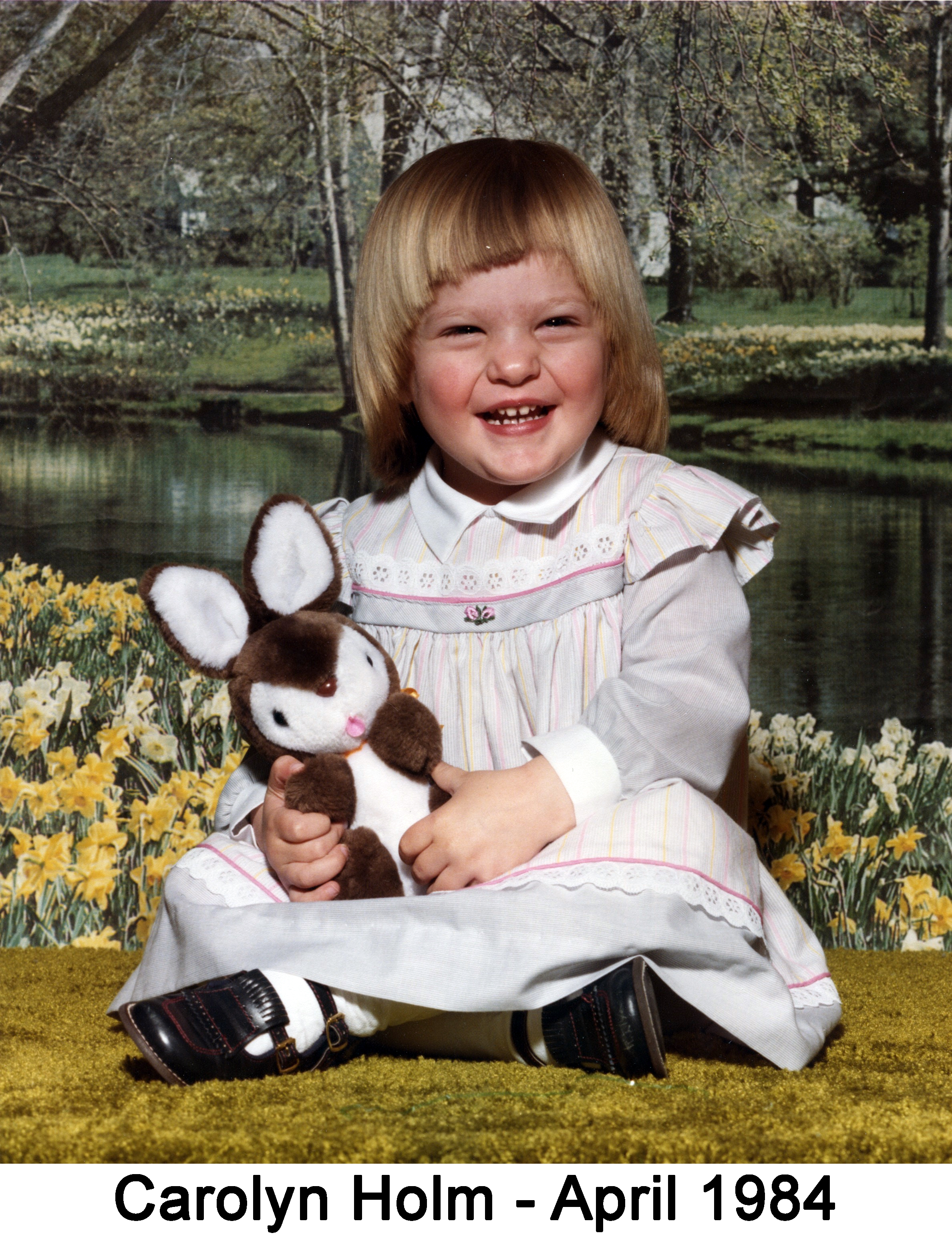 Carolyn Holm with a stuffed bunny is sitting in front of a phtograph of a stream and woods