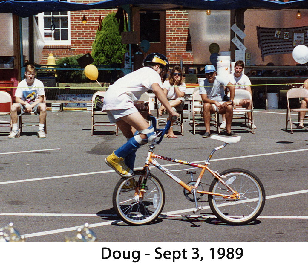 Doug is riding his orange bike, wearing a helmet and with his       head turned away from the camera. He is facing backwards and balancing        on the front wheel.