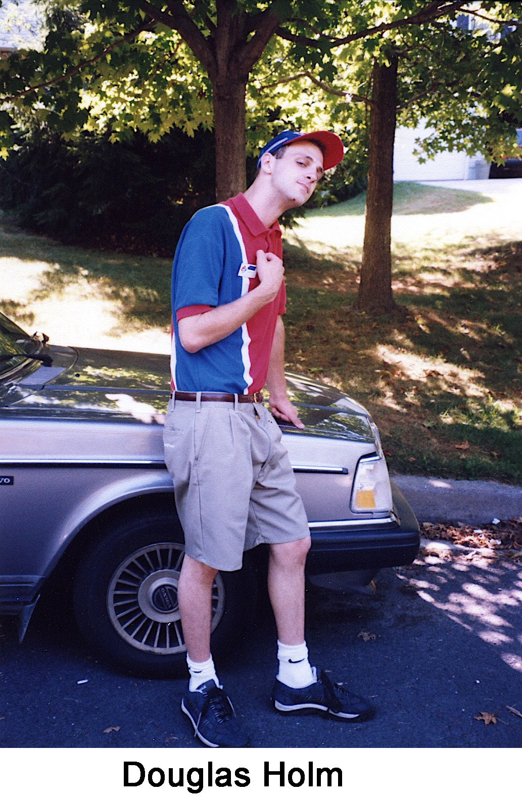 Doug is standing by the hood of the car, and wearing a red and blue shirt and hat     for his uniform.