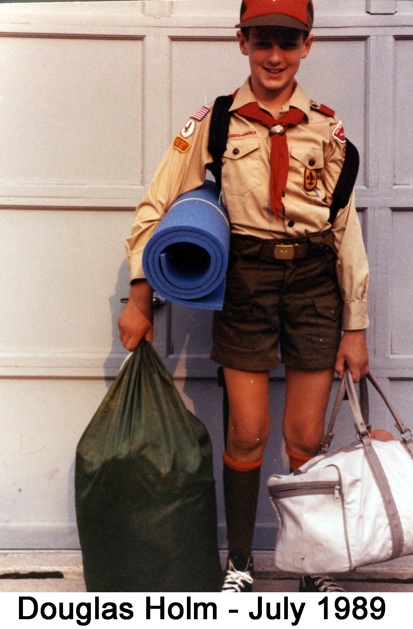 Douglas stands in front of our garage door in scout uniform with two bags 
          and a rolled-up foam mattress