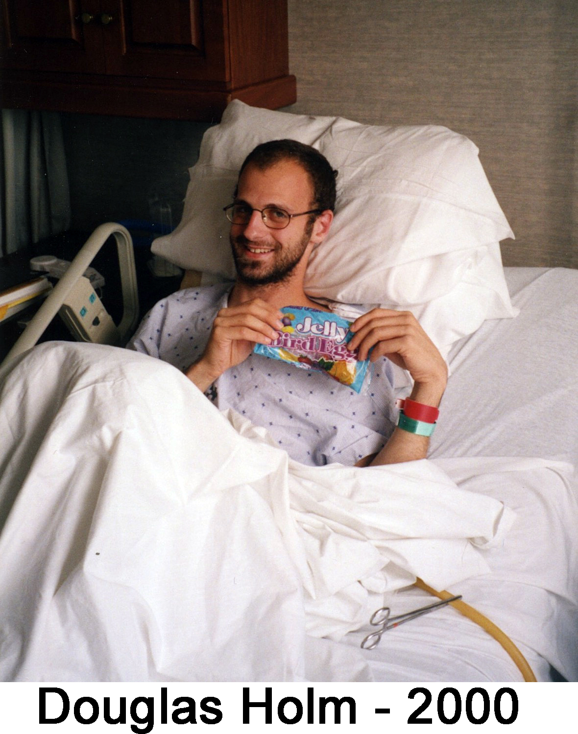 Doug Holm lying back on a pillow in a hospital bed and smiling with a bag of candy