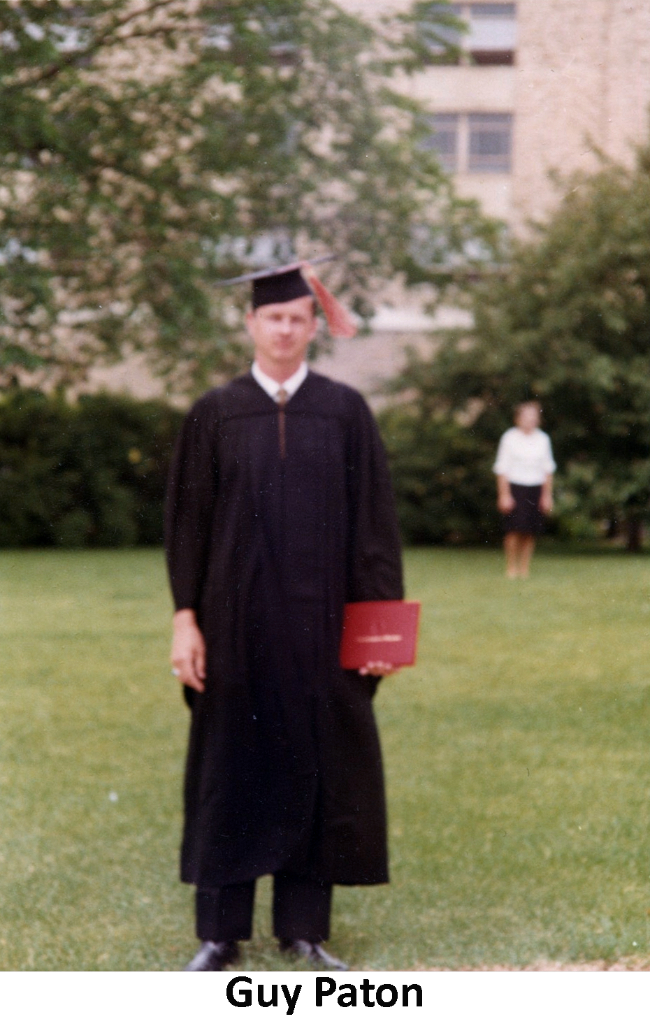 Guy Paton in his graduation robes standing on a lawn
              with trees and a tall building behind him