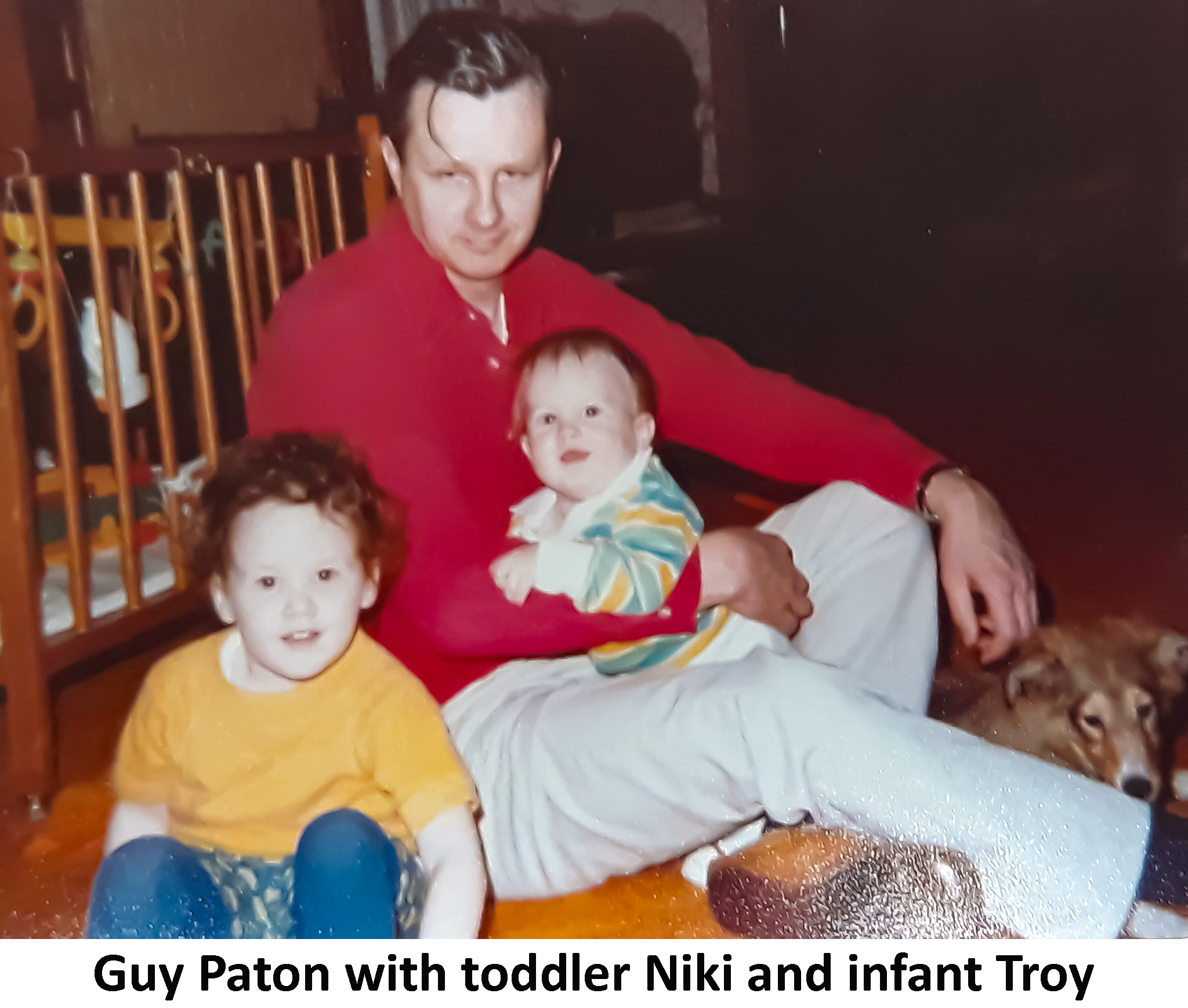 Guy Paton is sitting on the floor in front of a crib and holding Troy Paton.
                 Niki is sitting beside him.