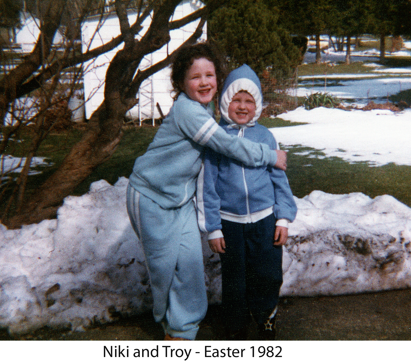 Niki holding Troy standing in their snowsuits in front of a snowbank.